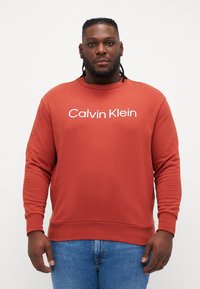 Ružovosfarbená mikina vyrobená z mäkkého materiálu, s okrúhlym výstrihom a bielym textom "Calvin Klein" cez hruď.