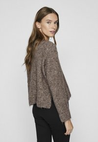 Brūns, pūkains adījums džemperis ar brīvu piegriezumu, kritiskām plecu šuvēm un īsu garumu. Valkāts ar melnām cieši pieguļošām biksēm.