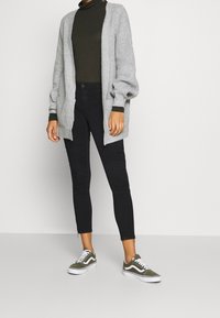 Cárdigan de punto gris claro con frente abierto, jersey de tortuga verde, jeans negros ajustados con detalles acanalados y zapatillas verde olivo con suelas blancas.