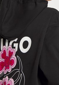 Sudadera negra con gran texto en blanco "HUGO" y un gráfico floral colorido en rosa y rojo en la parte trasera. Tejido suave, capucha ajustable.