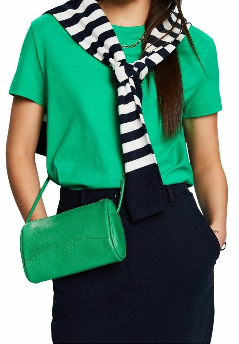 Personne portant une chemise vert vif, un pantalon noir, un pull rayé noir et blanc noué autour des épaules, et portant un petit sac à main vert.