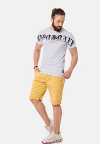 Weißes T-Shirt mit schwarzem Grafikdruck und Text, kombiniert mit gelben Shorts. Das Model trägt weiße Sportschuhe mit strukturierten Sohlen.
