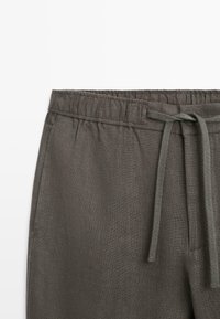 Gros plan sur un pantalon en lin gris avec taille élastique et cordon de serrage noué sur un fond blanc uni.