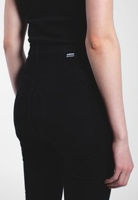 Jeans noirs taille haute en denim, avec des poches arrière, des détails de couture subtils et une étiquette de marque près de la ceinture.