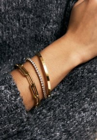 Tre braccialetti su un polso: un bracciale a catena d'oro, un sottile bracciale rigido d'oro e un braccialetto d'argento con pietre trasparenti, posizionati sopra un maglione grigio strutturato.
