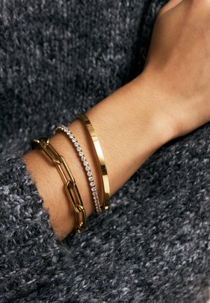 Drei Armbänder an einem Handgelenk: ein goldenes Gliederarmband, ein dünner goldener Armreif und ein silbernes Armband mit klaren Steinen, vor einem strukturierten grauen Pullover.