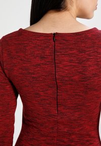 Vêtement rouge texturé à manches longues, avec un décolleté droit et une fermeture éclair verticale dans le dos. Le matériau semble extensible.