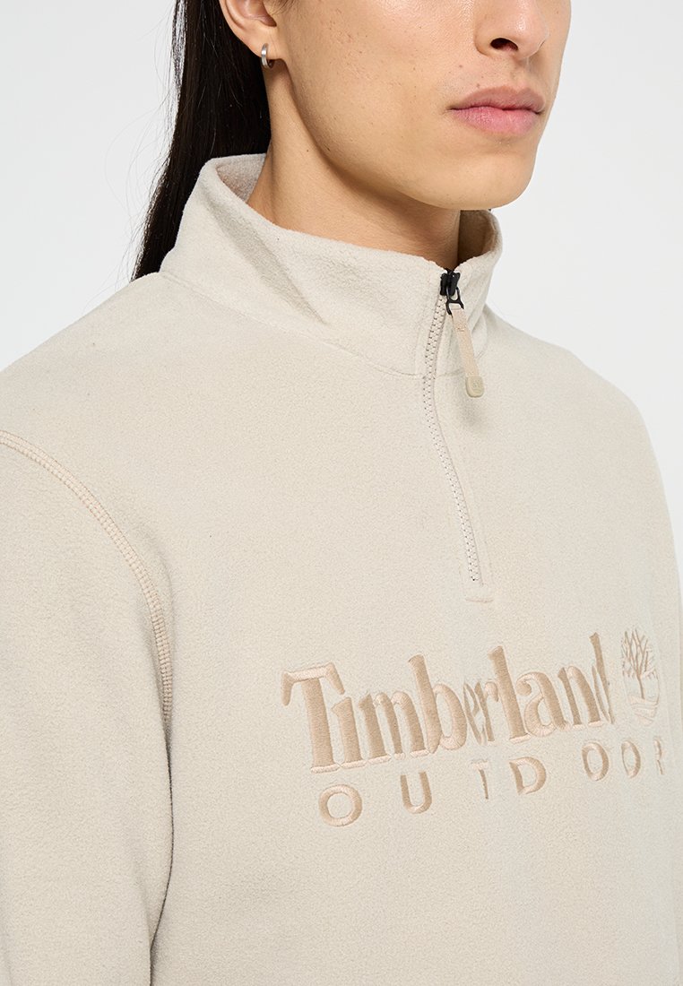 Pullover in pile color crema con colletto alto, chiusura a metà zip e logo "Timberland" in rilievo sul petto. Texture morbida, design minimal.