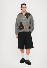 Suéter gris de punto con cremallera, adornado con un patrón de diamantes en beige y marrón. Combinado con shorts negros y zapatillas de tela negras bordadas.