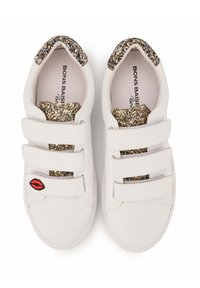 Baskets blanches avec trois sangles auto-agrippantes, un accent de talon pailleté doré et argenté, et une broderie de lèvres rouges sur la chaussure gauche.