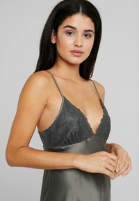 Chemise in satin di colore grigio scuro con dettagli in pizzo. Presenta un profondo scollo a V e spalline sottili. Texture liscia su tutta la superficie.