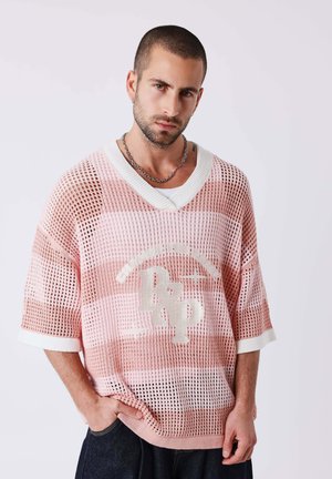 Uomo con capelli corti e barba indossa un maglione a rete rosa e bianco ampio con il logo "RXP", jeans scuri e una collana a catena d'argento.