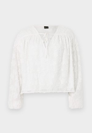 Witte korte blouse met lange mouwen, textuur van bloemige kant, V-halslijn en strikdetails aan de voorkant. Zacht, doorschijnend materiaal.