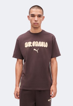 Jeune homme portant un t-shirt marron St. Pauli Puma et un short assorti, debout devant un fond uni gris clair.