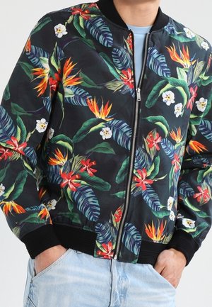 Schwarze Bomberjacke mit buntem tropischem Blumenmuster, getragen über einem hellblauen Hemd, kombiniert mit hellblauen Jeans.