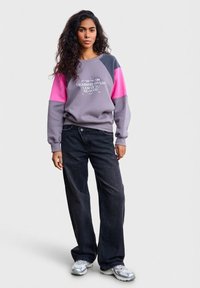 Šedá mikina s růžovými a modrými rukávy, s potiskem textu. Kombinována s černými širokými džínami a šedými teniskami.