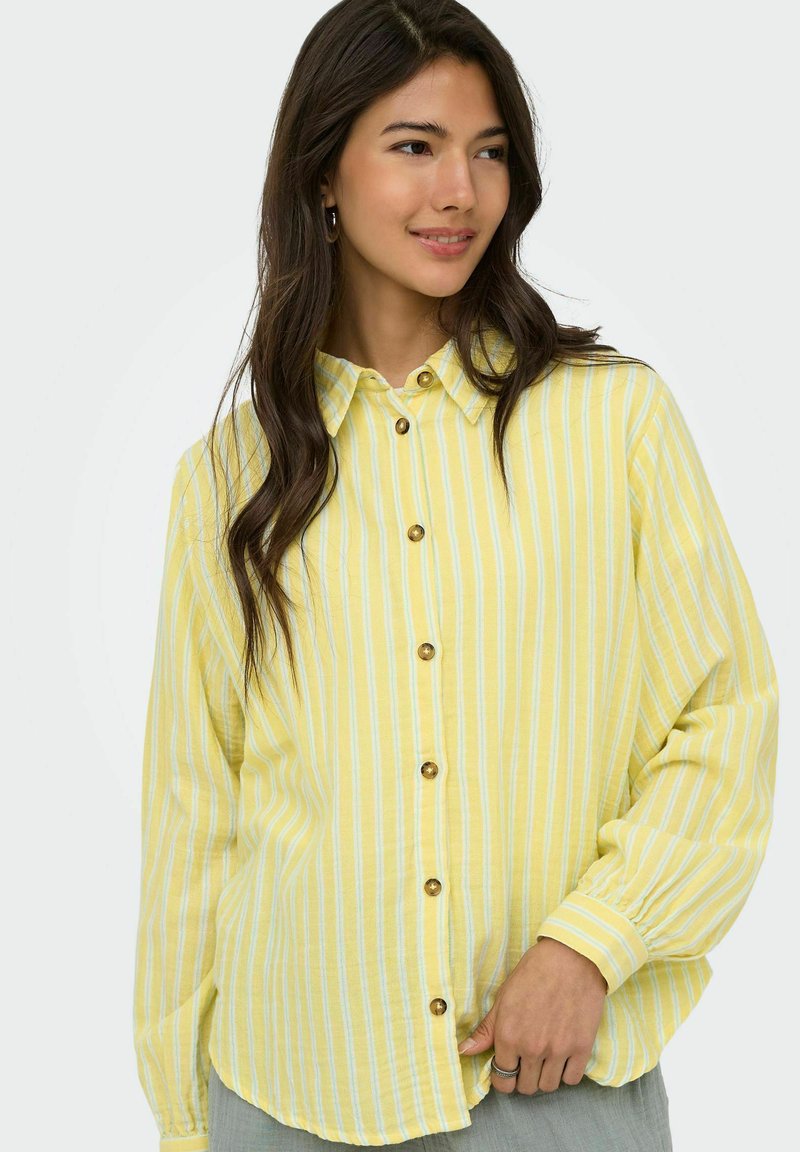 Jeune femme aux longs cheveux foncés, portant une chemise boutonnée rayée jaune et blanche et un pantalon gris clair, regardant sur le côté avec un léger sourire.