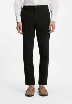 Mann trägt schwarze, maßgeschneiderte Hose, weißes Hemd und hellbraune Loafers, steht vor einem schlichten weißen Hintergrund.