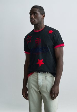 Ung mand i sort T-shirt med røde stjerner og symboler, lag på en rød skjorte, iført lyse bukser, står mod en ensfarvet baggrund.