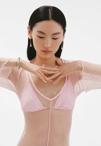 Top de malla rosa de manga larga con un escote en V profundo y textura translúcida, superpuesto sobre un sujetador rosa claro. Joyas plateadas que acentúan las manos.