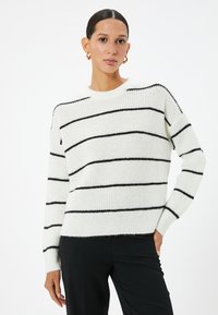 Pull rayé en tissu tricoté doux ; blanc avec des rayures noires. Il dispose d'un col rond et de manches longues avec des poignets côtelés.