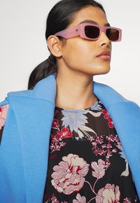 Óculos de sol retangulares rosa claro com lentes escuras, complementados por uma camada exterior azul com colarinho sobre uma blusa de padrão floral com fundo preto e cores vivas.