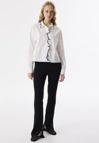 Camisa blanca de manga larga con ribete de volantes negros a lo largo de la abertura de botones, combinada con pantalones negros ajustados y zapatos planos negros.