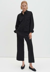 Camicia nera intrecciata con colletto e chiusura frontale con bottoni, abbinata a pantaloni neri a gamba larga. Calzature nere, piatte, con una texture arricciata.