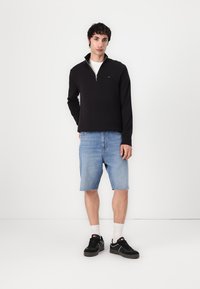 Pull-over noir à fermeture éclair, shorts en denim bleu clair, baskets noires avec semelles blanches, et chaussettes blanches montant jusqu'à la cheville. Tenue simple et décontractée.