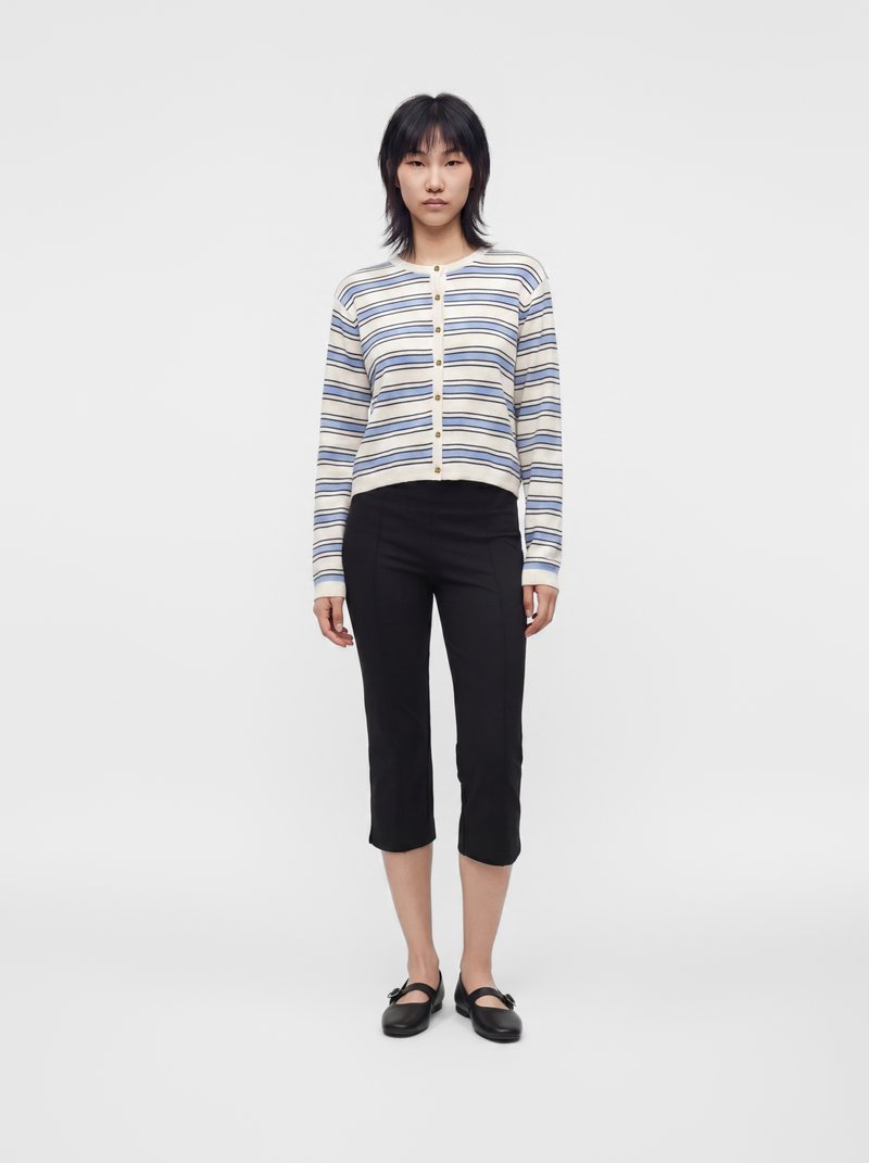 Personne debout face à l'avant, portant un cardigan rayé à manches longues, un pantalon noir court et des chaussures noires sur un fond blanc uni.