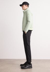 Jachetă matlasată verde deschis cu guler înalt, buzunare laterale și închidere cu fermoar. Asortată cu pantaloni negri și pantofi negri cu șiret.