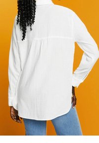 Chemise blanche texturée à manches longues avec un col boutonné, un ourlet arrondi et un pli au dos, associée à un jean bleu clair sur un fond orange.