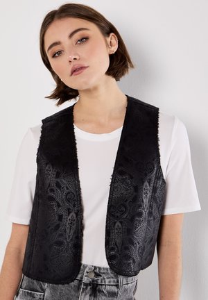 Jonge vrouw met kort bruin haar draagt een wit T-shirt, een zwart met patroon vest en grijs acid-wash jeans, poserend tegen een witte muur.