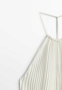 Robe blanche plissée sans manches avec fines bretelles spaghetti et un col froncé, suspendue sur un fond blanc.