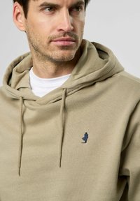 Beige hoodie met een trekkoordcapuchon, voorzien van een klein blauw geborduurd logo. Gemaakt van zacht materiaal met een getextureerde afwerking.