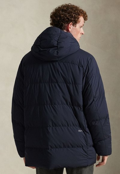 Tamsiai mėlyna pūsta striukė su kapišonu, pasižyminti siuvinėtais skyriais ir laisvu siluetu. Švelnus logotipo detalės prigludimas prie apatinės dalies, minkšta tekstūra.