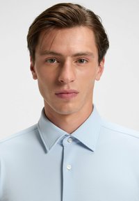 Hellblauses Polohemd mit strukturiertem Stoff, spitzem Kragen und einer Drei-Knopf-Leiste, getragen von einem Modell mit kurzer Frisur.