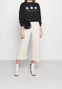Felpa nera con margherite ricamate e testo, pantaloni ampi crema, borsa a tracolla nera e stivaletti bianchi con dettagli neri.