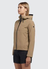 Giacca a cappuccio beige con zip frontale, accenti neri e un logo sul petto. Il materiale sembra liscio e resistente all'acqua.