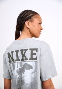 Szara bawełniana koszulka z krótkim rękawem, z dużym czarnym logo "NIKE" oraz grafiką Skrzydlatej Zwycięstwa z Samotraki na plecach.