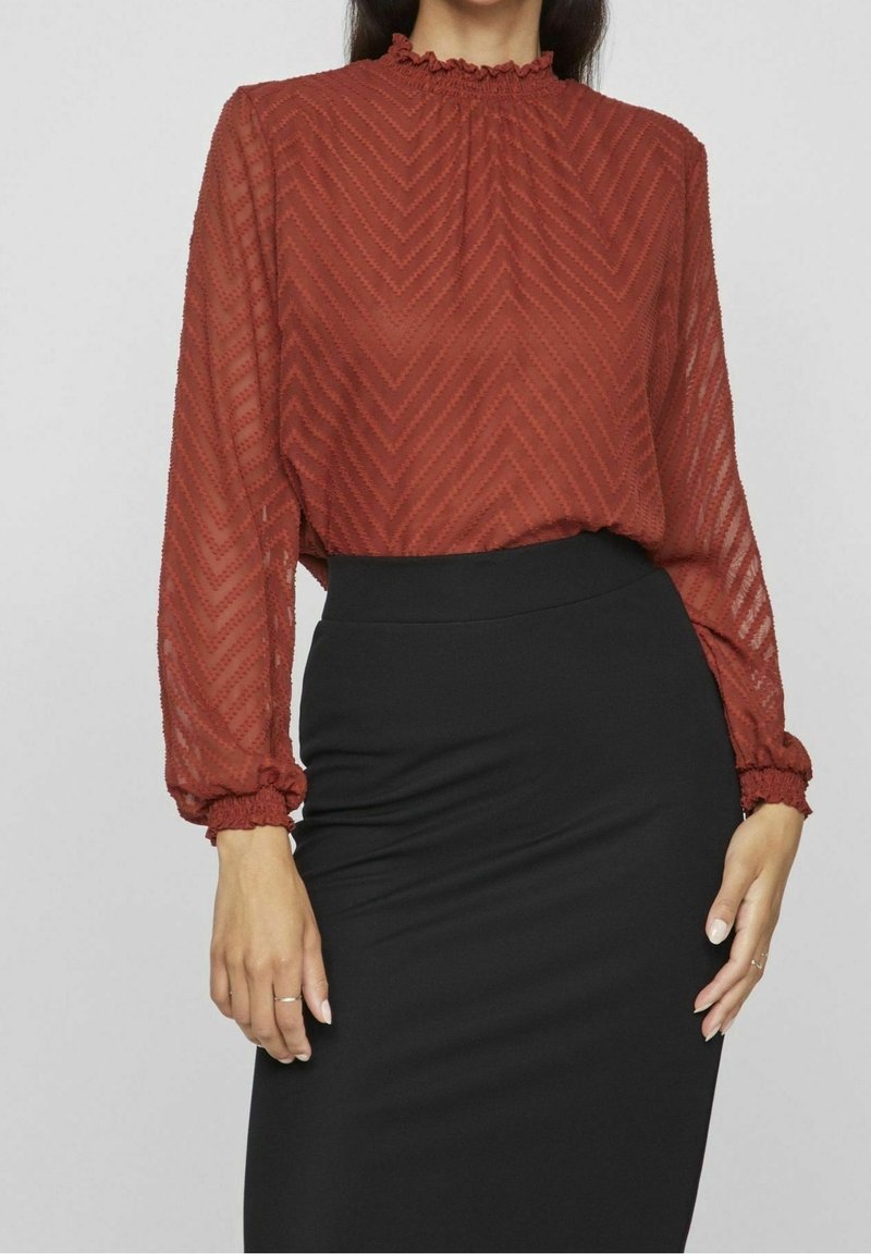 Femme portant un chemisier transparent de couleur rouille avec un motif chevron et un col montant, associé à une jupe noire ajustée.
