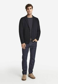 Svart kardiagan med slagkrage, kombinerad med en grå skjorta och mörka jeans, avslutad med beige avslappnade skor. Enkel knappdesign.