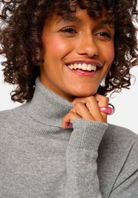 Pull à col roulé gris avec une texture côtelée et des manches longues. Le modèle porte une bague rose, affichant une expression souriante et des cheveux bouclés.
