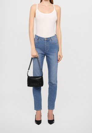 Mujer con top blanco sin mangas, jeans azules de talle alto, tacones negros puntiagudos, sosteniendo un bolso pequeño negro con la etiqueta "LOVE MOSCHINO".