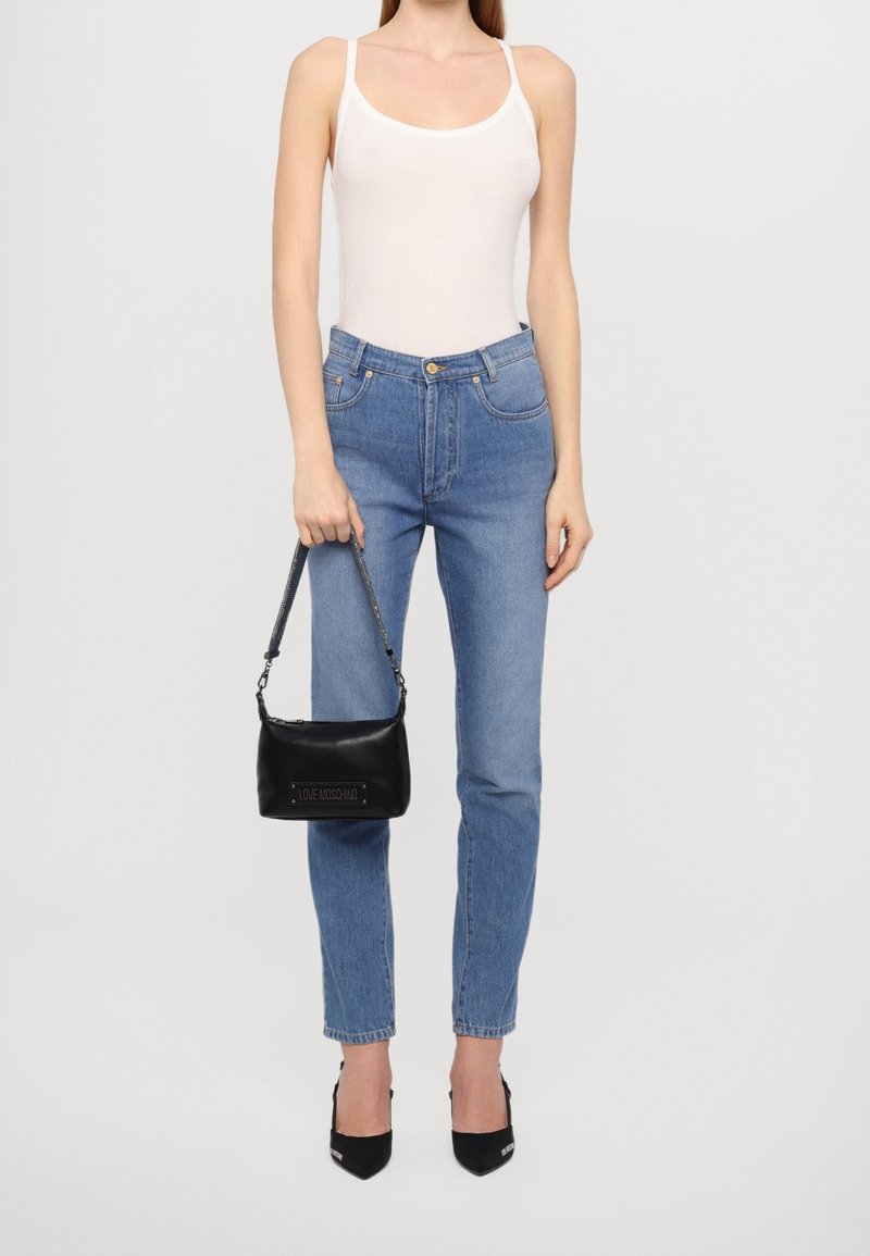 Femme portant un top blanc sans manches, un jean taille haute bleu, des talons pointus noirs, tenant un petit sac à main noir avec l'étiquette "LOVE MOSCHINO".