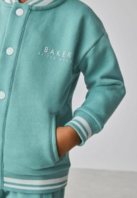 Zelenomodrá mikina s kapucňou, bielymi gombíkmi a logom "BAKER by TED BAKER." Má pruhované manžety a dve bočné vrecká.