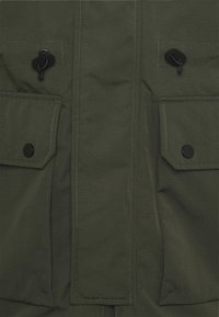 Framsidan av en olivgrön jacka med två fickor med lock som stängs med svarta tryckknappar och snoddar ovanför varje ficka.