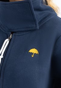 Felpa con zip color blu navy e colletto alto, con un piccolo ricamo di un ombrello giallo sul petto e dettagli con cordini bianchi. Texture morbida.