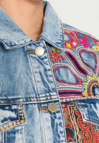 Džínová bunda se světle modrým omakem, zdobená barevným vyšíváním v abstraktních vzorech, kovovým knoflíkem a kontrastními švy.