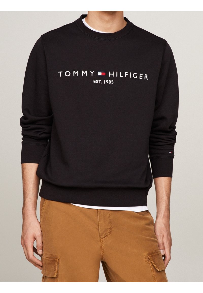 Homme portant un sweat-shirt noir Tommy Hilfiger avec le logo et "EST. 1985", assorti à un pantalon cargo marron et un sous-vêtement blanc.